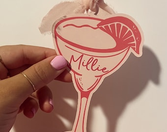 May include: A flat, die-cut ornament shaped like a cocktail glass with a pink and white color scheme. The glass features a lime wedge garnish and the name Millie printed in a script font. A light pink fabric ribbon is attached to the top loop.