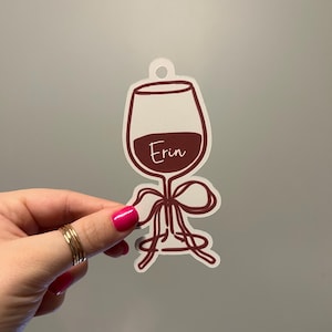 Puede incluir: Un adorno en forma de copa de vino con un relleno de vino color burdeos y el nombre "Erin" en fuente cursiva. El adorno tiene un contorno blanco y un lazo decorativo. Lo sostiene una mano con esmalte de uñas rosa y anillos dorados.