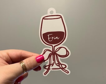 May include: A die-cut sticker shaped like a wine glass with a dark red outline and filled with dark red liquid. The name Erin is written in white script across the center of the glass. A dark red ribbon is tied around the stem of the wine glass.