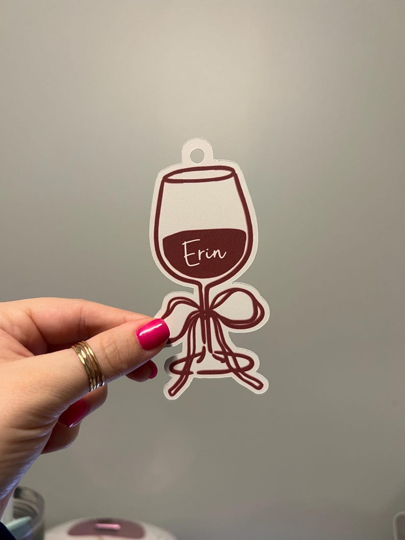 May include: A die-cut sticker shaped like a wine glass with a dark red outline and filled with dark red liquid. The name Erin is written in white script across the center of the glass. A dark red ribbon is tied around the stem of the wine glass.