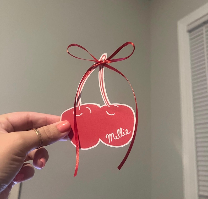 May include: A red cherry-shaped paper cutout with white accents and the name "Millie" in cursive. A red ribbon is attached at the top, forming a loop and bow. The cutout is held up against a gray background.