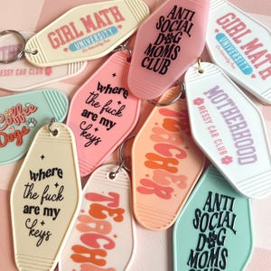 May include: A collection of colorful keychains with various phrases and designs. The keychains are shaped like motel keys and feature phrases like "Girl Math University", "Anti Social Dog Moms Club", "Where the fuck are my keys", and "Teacher".