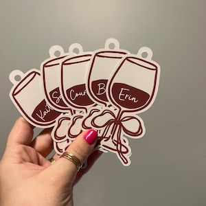 May include: A set of five die-cut wine glass tags featuring a dark red wine graphic, a decorative bow at the stem, and a circular hole at the top for hanging. Each tag is personalized with a white script name including Kait, Courtney, and Erin.