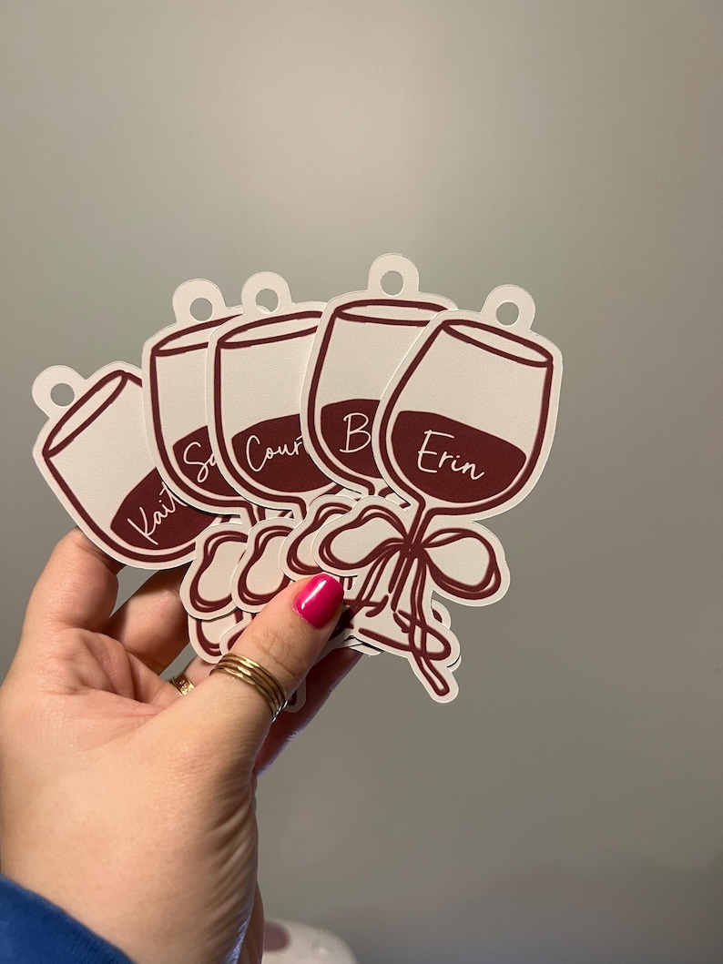 May include: A set of five die-cut wine glass tags featuring a dark red wine graphic, a decorative bow at the stem, and a circular hole at the top for hanging. Each tag is personalized with a white script name including Kait, Courtney, and Erin.