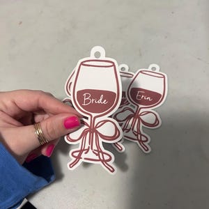 May include: A set of die-cut wine glass tags featuring a maroon outline and a maroon bow at the base. The tags are white with a maroon-colored wine graphic. One tag displays the word Bride in white script, and another displays the name Erin.