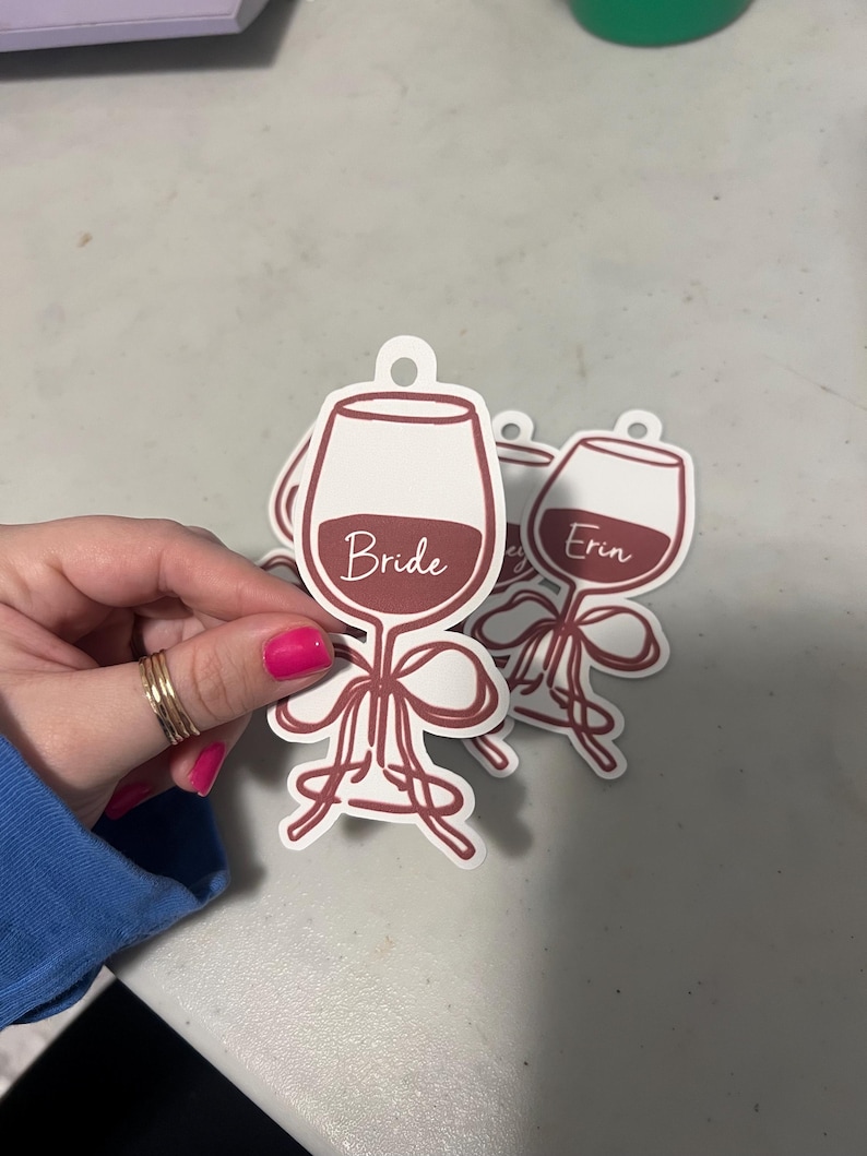May include: A set of die-cut wine glass tags featuring a maroon outline and a maroon bow at the base. The tags are white with a maroon-colored wine graphic. One tag displays the word Bride in white script, and another displays the name Erin.