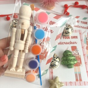 以下が含まれることがあります： ペイントとブラシが付いた木製くるみ割り人形キット。キットには、ピンク、ブルー、オレンジ、パープル、ピーチの5つのペイントポットが含まれています。パッケージには「あなた自身のくるみ割り人形をペイントしましょう」というテキストが書かれています。