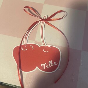 May include: A red cherry-shaped tag with the name "Millie" in white script. The tag is adorned with a red ribbon tied in a bow at the top. The background is a pink and white checkered pattern.