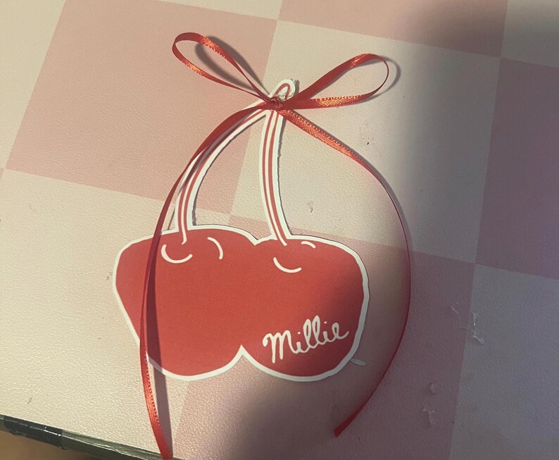 May include: A red cherry-shaped tag with the name "Millie" in white script. The tag is adorned with a red ribbon tied in a bow at the top. The background is a pink and white checkered pattern.
