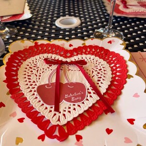 May include: A Valentine's Day themed place setting featuring a heart-shaped doily with a red ribbon and a cherry design. The doily is layered over a white plate with gold trim and scattered heart confetti.