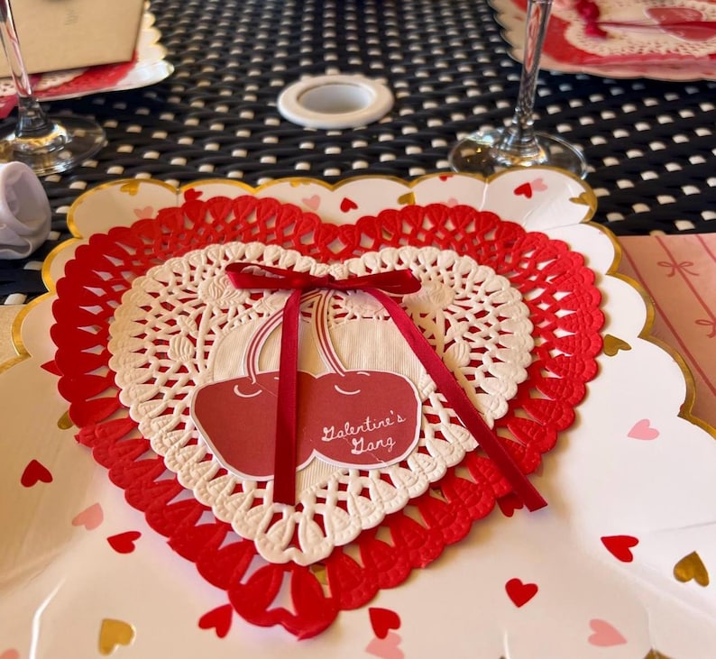 May include: A Valentine's Day themed place setting featuring a heart-shaped doily with a red ribbon and a cherry design. The doily is layered over a white plate with gold trim and scattered heart confetti.