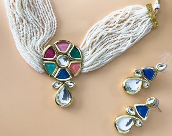 May include: A white pearl necklace with a multi-colored pendant and matching earrings. The pendant features a round design with pink, green, blue, and clear stones. The earrings are teardrop shaped with blue and clear stones.