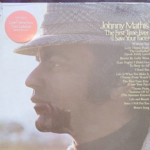 May include: A vintage vinyl record album cover featuring a black and white photo of Johnny Mathis. The album title is "The First Time Ever (I Saw Your Face)" and lists the songs included on the record. The album cover is a light brown color with a white border.