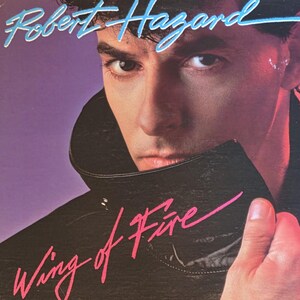 May include: Album cover for Robert Hazard's "Wing of Fire." The cover features a man with dark hair and a leather jacket, with the album title in pink script and the artist's name in blue script. The background is a gradient of purple and red.