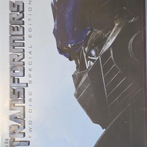 Op de afbeelding: Een dvd-hoes voor de film "Transformers: Two-Disc Special Edition". De voorkant toont een close-up van een robotgezicht in blauwe, grijze en zilveren tinten. De titel staat in grote, zilveren letters.
