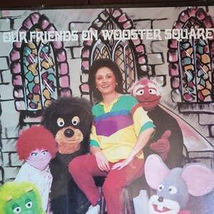 Puede incluir: Un grupo de títeres y una mujer con una camisa colorida se encuentran frente a una pared de ladrillo con ventanas de vidrieras. El texto "Our Friends on Wooster Square" está en la parte superior de la imagen.