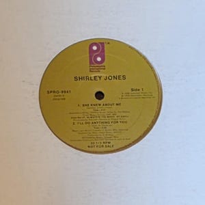 Puede incluir: Un álbum de vinilo con una etiqueta amarilla con el texto "SHIRLEY JONES". La portada del álbum es blanca. La etiqueta incluye títulos de canciones como "SHE KNEW ABOUT ME" y "I'LL DO ANYTHING FOR YOU".