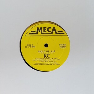 Könnte beinhalten: Eine Schallplatte mit einem gelben Label mit dem Text "GIVE IT UP 5:18" und den Initialen des Künstlers "KC". Die Schallplatte befindet sich in einer weißen Papierhülle. Das Label enthält auch das "MECA"-Logo.
