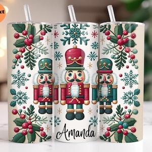 May include: A white tumbler with a green, red, and gold design featuring a nutcracker. The tumbler has the name "Amanda" printed on the bottom.