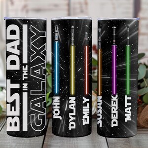 K&ouml;nnte beinhalten: Drei schwarze, isolierte Becher mit Weltraum-Motiv. Auf einem Becher steht in Wei&szlig; "BEST DAD IN THE GALAXY". Die anderen beiden Becher haben farbenfrohe Lichtschwert-Designs und die Namen: John, Dylan, Emily, Susan, Derek und Matt.