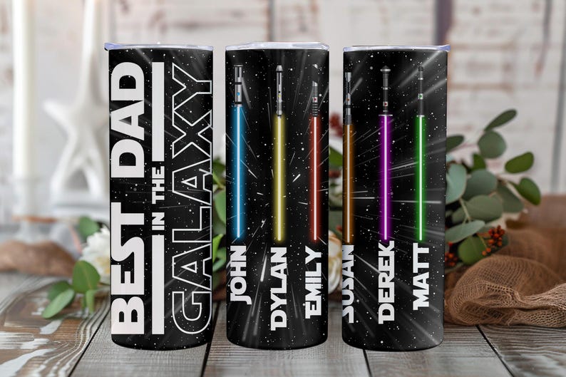 K&ouml;nnte beinhalten: Drei schwarze, isolierte Becher mit Weltraum-Motiv. Auf einem Becher steht in Wei&szlig; "BEST DAD IN THE GALAXY". Die anderen beiden Becher haben farbenfrohe Lichtschwert-Designs und die Namen: John, Dylan, Emily, Susan, Derek und Matt.