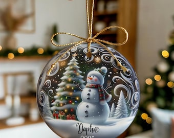 May include: A round ceramic Christmas ornament featuring a snowman wearing a plaid scarf, a decorated pine tree, and swirling white patterns on a dark background. The name Daphne and the year 2025 are printed in black script. It hangs from a gold cord.