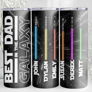 K&ouml;nnte beinhalten: Ein schwarzer Becher mit dem Text "BEST DAD in the GALAXY" in Wei&szlig;. Der Becher zeigt farbenfrohe Lichtschwert-Designs mit den Namen: John, Dylan, Emily, Susan, Derek und Matt. Das Design ist vor einem Sternenhintergrund.