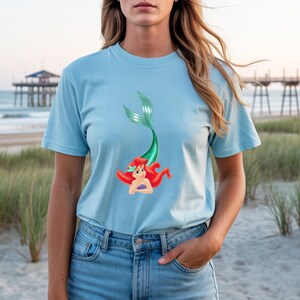 May include: Light blue t-shirt featuring a cartoon mermaid with red hair and a green tail. The mermaid is smiling and has a purple seashell bra. The shirt is made of a soft material and is perfect for casual wear.