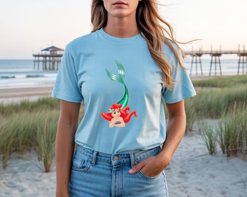 May include: Light blue t-shirt featuring a cartoon mermaid with red hair and a green tail. The mermaid is smiling and has a purple seashell bra. The shirt is made of a soft material and is perfect for casual wear.