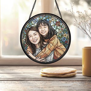 May include: Round stained glass art piece featuring two smiling people embracing, set against a floral background. The artwork is framed in black and hangs from a chain. The scene is set on a wooden surface with a stack of pancakes and a yellow mug.