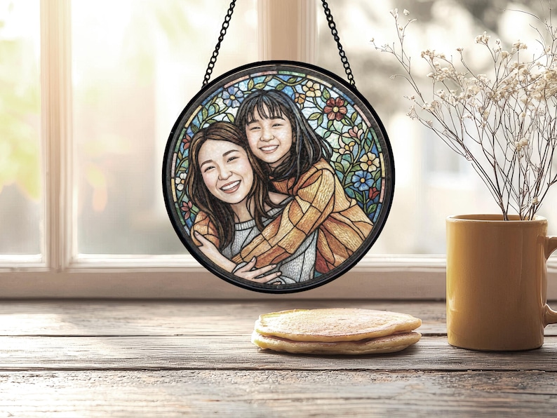 May include: Round stained glass art piece featuring two smiling people embracing, set against a floral background. The artwork is framed in black and hangs from a chain. The scene is set on a wooden surface with a stack of pancakes and a yellow mug.