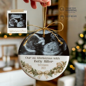 May include: A round ceramic ornament featuring a black and white ultrasound image at the top. The bottom section displays the text "Our 1st Christmas with Baby MILLER" and "SEPTEMBER 2027" above a decorative illustration of baby shoes, a teddy bear, and a bow.