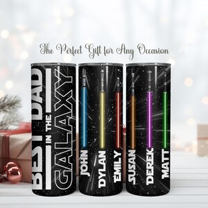 K&ouml;nnte beinhalten: Drei schwarze isolierte Becher mit Weltraum-Motiv. Auf einem Becher steht "BEST DAD IN THE GALAXY" mit einem Lichtschwert-Design. Die anderen beiden Becher haben Lichtschwerter in Blau, Gelb, Rot, Lila und Gr&uuml;n mit Namen.