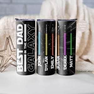 K&ouml;nnte beinhalten: Schwarze isolierte Becher mit Weltraum-Motiv. Auf einem Becher steht "BEST DAD IN THE GALAXY". Die anderen Becher zeigen farbenfrohe Lichtschwert-Designs mit den Namen: John, Dylan, Emily, Susan, Derek und Matt.
