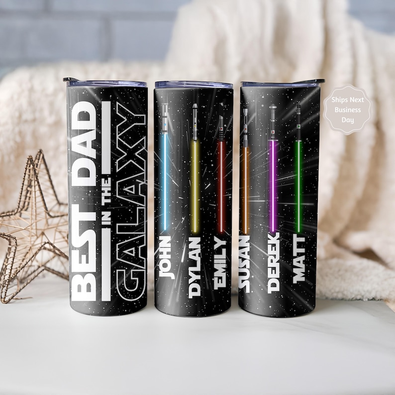 K&ouml;nnte beinhalten: Schwarze isolierte Becher mit Weltraum-Motiv. Auf einem Becher steht "BEST DAD IN THE GALAXY". Die anderen Becher zeigen farbenfrohe Lichtschwert-Designs mit den Namen: John, Dylan, Emily, Susan, Derek und Matt.