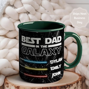 May include: A black ceramic mug with a green handle and rim. The mug features the text "BEST DAD in the GALAXY" in white, with the names Dylan, Emily, and John below. The design includes colorful light streaks.