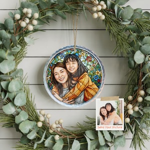 May include: A round, stained-glass style ornament featuring a portrait of two people hugging, surrounded by colorful flowers. The ornament hangs from a gold string, set against a green wreath with white berries. A small photo with the text "Send Your Picture" is also visible.