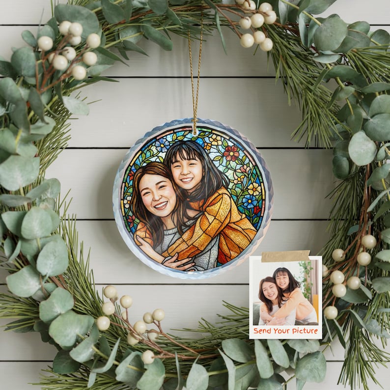 May include: A round, stained-glass style ornament featuring a portrait of two people hugging, surrounded by colorful flowers. The ornament hangs from a gold string, set against a green wreath with white berries. A small photo with the text "Send Your Picture" is also visible.