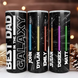 K&ouml;nnte beinhalten: Drei schwarze, isolierte Becher mit Weltraumthema. Jeder Becher hat ein unterschiedlich farbiges Lichtschwert und einen Namen. Der Text "BEST DAD IN THE GALAXY" ist auf dem ersten Becher zu sehen.