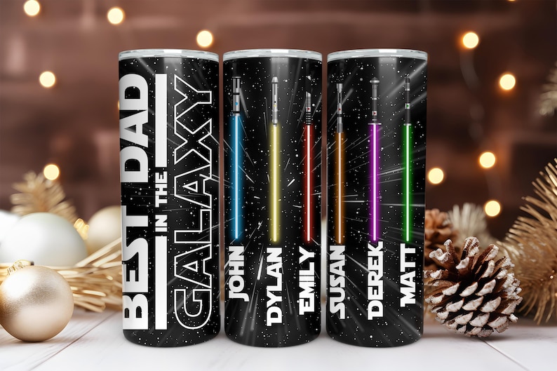 K&ouml;nnte beinhalten: Drei schwarze, isolierte Becher mit Weltraumthema. Jeder Becher hat ein unterschiedlich farbiges Lichtschwert und einen Namen. Der Text "BEST DAD IN THE GALAXY" ist auf dem ersten Becher zu sehen.