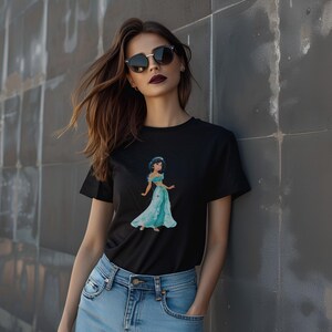 May include: Black t-shirt featuring a watercolor illustration of a princess in a teal gown. The graphic tee has short sleeves and a crew neck. The model is wearing blue jeans and sunglasses.
