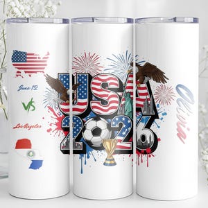 Może przedstawiać: Biały kubek termiczny z patriotycznym wzorem. Zawiera napis "USA 2026" z piłką nożną, orłem i fajerwerkami. Zawiera mapę USA i tekst "June 12 vs Los Angeles".