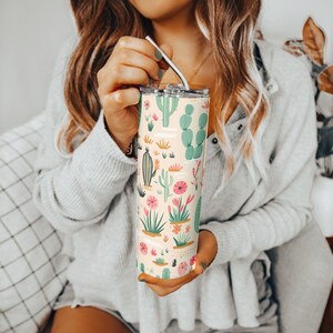 May include: A tall, cylindrical stainless steel tumbler featuring a light cream background with a repeating pattern of various green cacti and pink desert flowers, topped with a clear lid and a thin, curved silver metal straw for drinking.