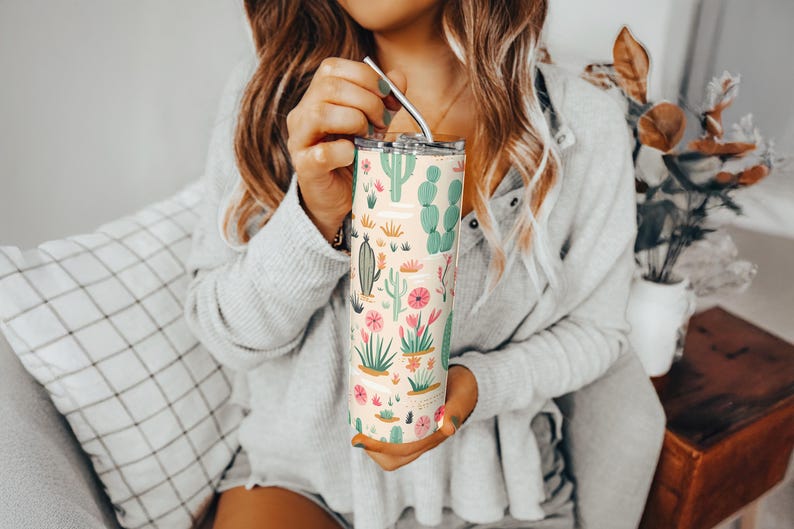 May include: A tall, cylindrical stainless steel tumbler featuring a light cream background with a repeating pattern of various green cacti and pink desert flowers, topped with a clear lid and a thin, curved silver metal straw for drinking.