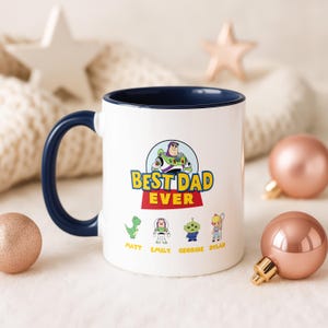 May include: A white ceramic mug with a dark blue handle and interior rim. The front features a graphic of Buzz Lightyear above the text "BEST DAD EVER" in yellow and red, with four small character icons labeled Matt, Emily, Georgie, and Dylan.