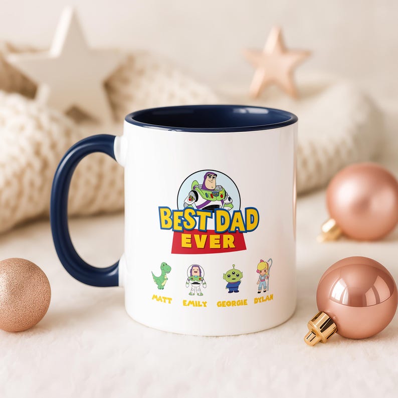 May include: A white ceramic mug with a dark blue handle and interior rim. The front features a graphic of Buzz Lightyear above the text "BEST DAD EVER" in yellow and red, with four small character icons labeled Matt, Emily, Georgie, and Dylan.