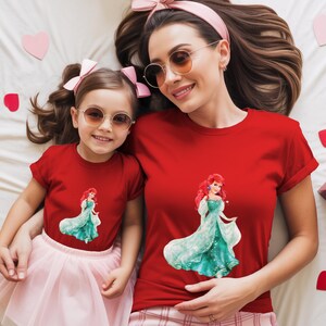 May include: Two red t-shirts, one adult and one child size, featuring a graphic of a princess in a teal gown. The adult is wearing sunglasses and a pink headband. The child is wearing a pink skirt and sunglasses. Pink hearts and gift boxes are scattered around.