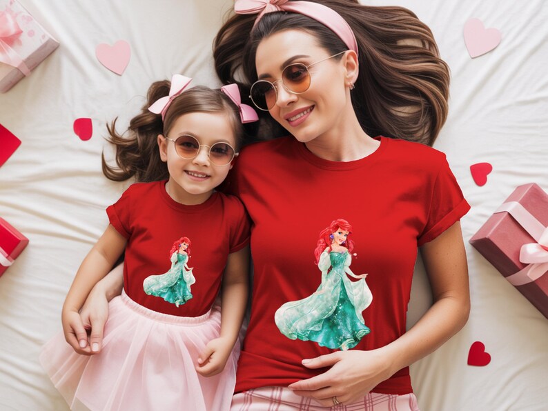 May include: Two red t-shirts, one adult and one child size, featuring a graphic of a princess in a teal gown. The adult is wearing sunglasses and a pink headband. The child is wearing a pink skirt and sunglasses. Pink hearts and gift boxes are scattered around.