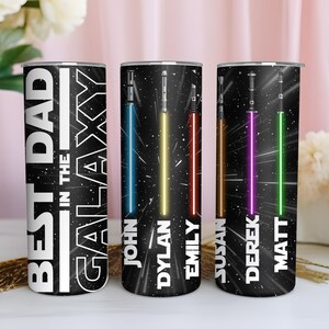 K&ouml;nnte beinhalten: Drei schwarze isolierte Becher mit Weltraum-Motiv. Auf einem Becher steht in Wei&szlig; "BEST DAD IN THE GALAXY". Die anderen beiden Becher zeigen Lichtschwerter in Blau, Gelb, Rot, Lila und Gr&uuml;n mit Namen darunter. Ein dekorativer Artikel f&uuml;r einen Star Wars-Fan.