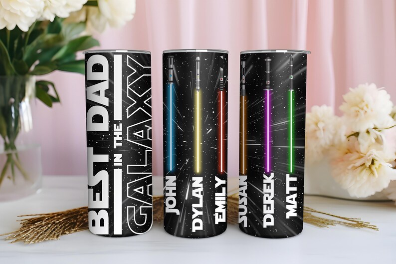 K&ouml;nnte beinhalten: Drei schwarze isolierte Becher mit Weltraum-Motiv. Auf einem Becher steht in Wei&szlig; "BEST DAD IN THE GALAXY". Die anderen beiden Becher zeigen Lichtschwerter in Blau, Gelb, Rot, Lila und Gr&uuml;n mit Namen darunter. Ein dekorativer Artikel f&uuml;r einen Star Wars-Fan.